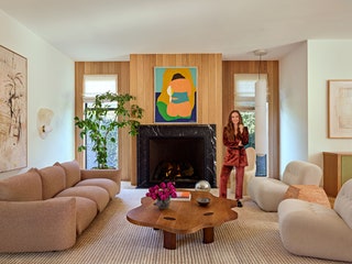 Ariel Kaye in burgundy suit standing in living area in front of fireplace flanked by tall windows wooden coffee table...