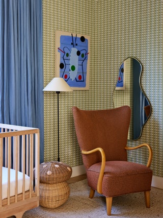Corner of nursery covered in green patterned wallpaper blue abstract artwork hanging on wall above rust colored chair crib