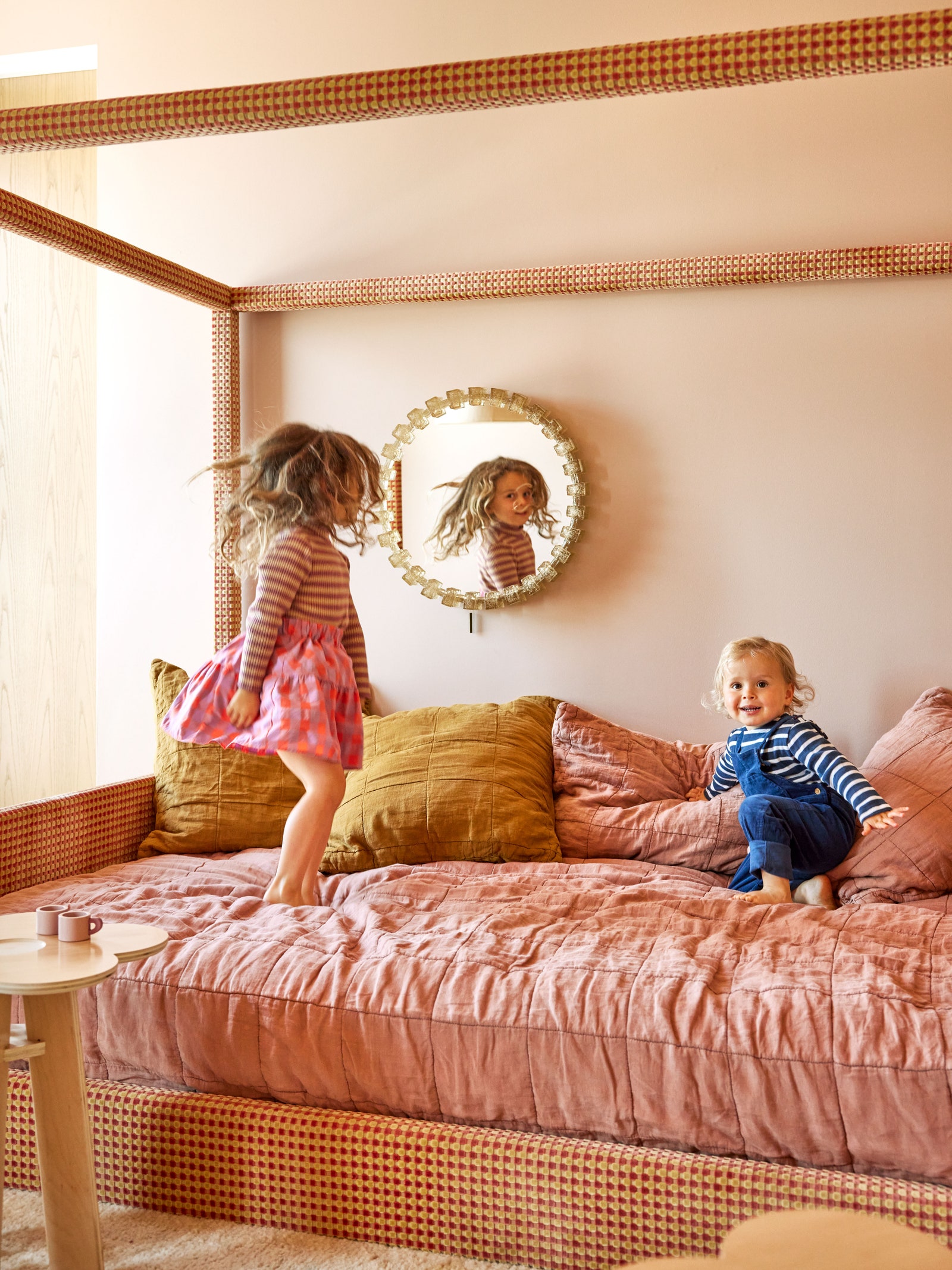 Two young children bouncing on a pink bed small round mirror on wall